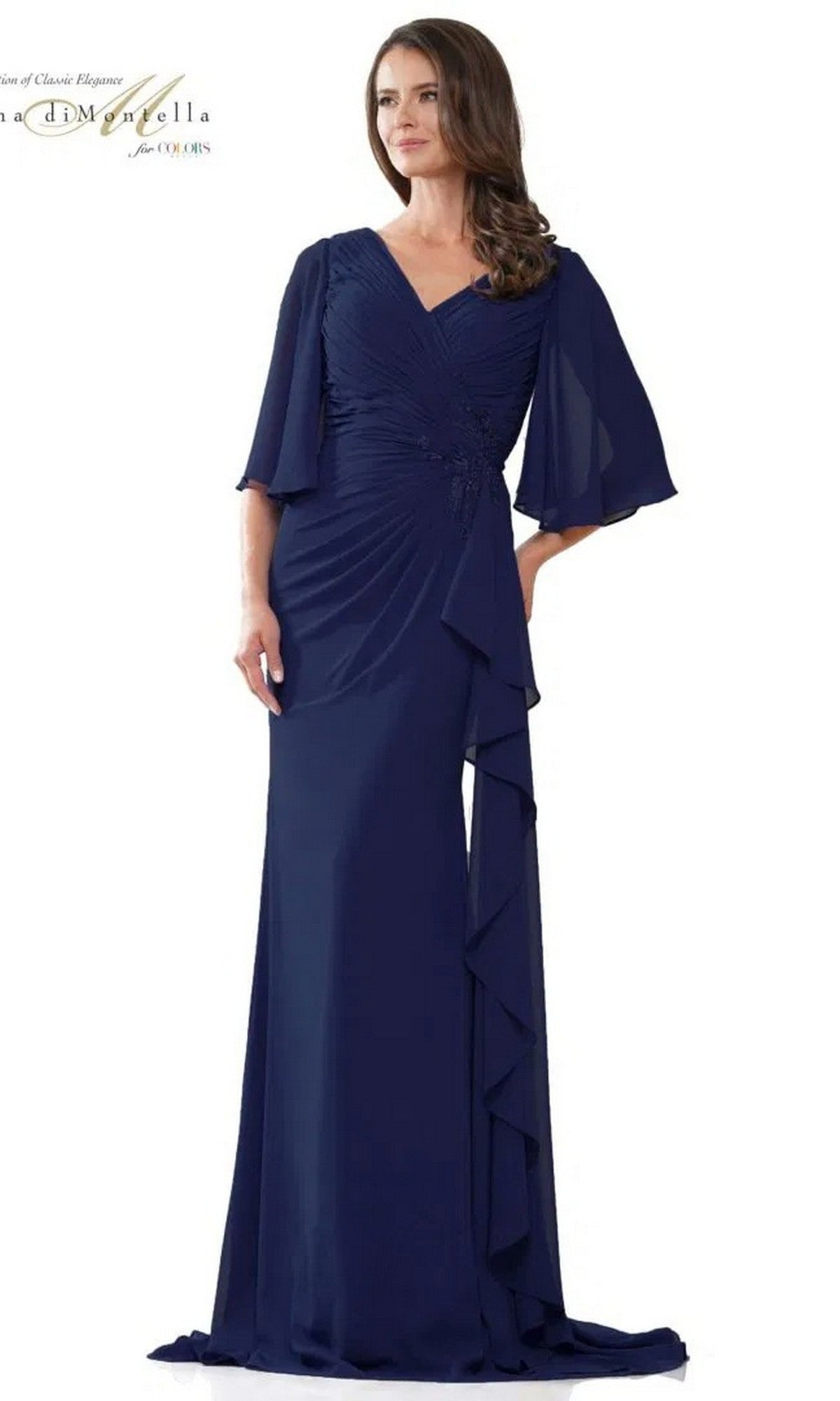 A woman stands against a plain background in the Rina Di Montella RD2935 long formal dress in navy, featuring chiffon fabric, a ruched bodice, draped details, and flutter sleeves.